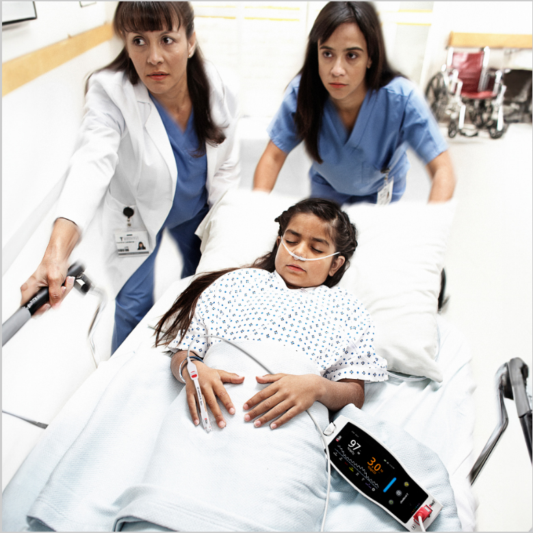 A child patient wearing a Masimo sensor attached to Masimo device on a gurney being pushed by two clinicians down a hospital corridor. 